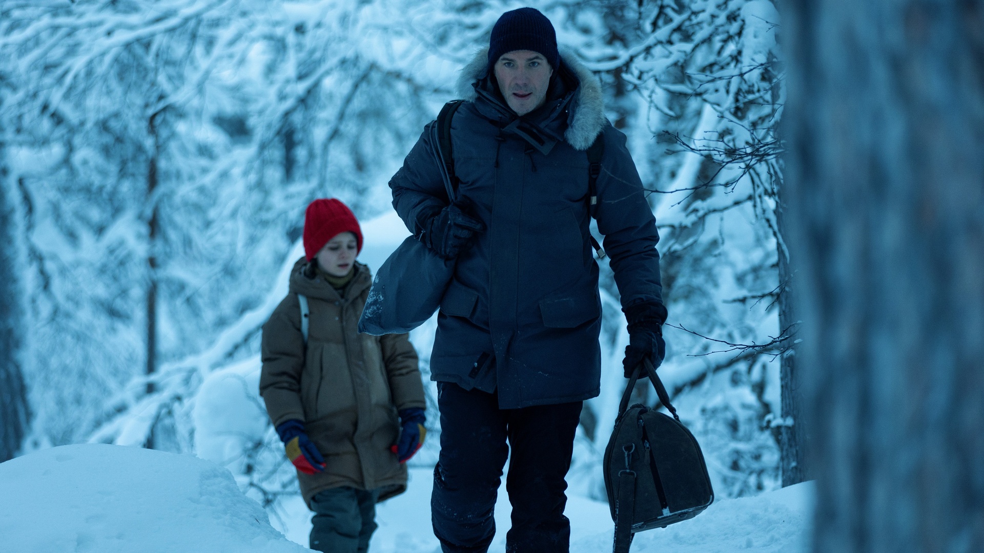 A young girl and her father are wearing thick winter clothing as they traverse through a snowy forest.