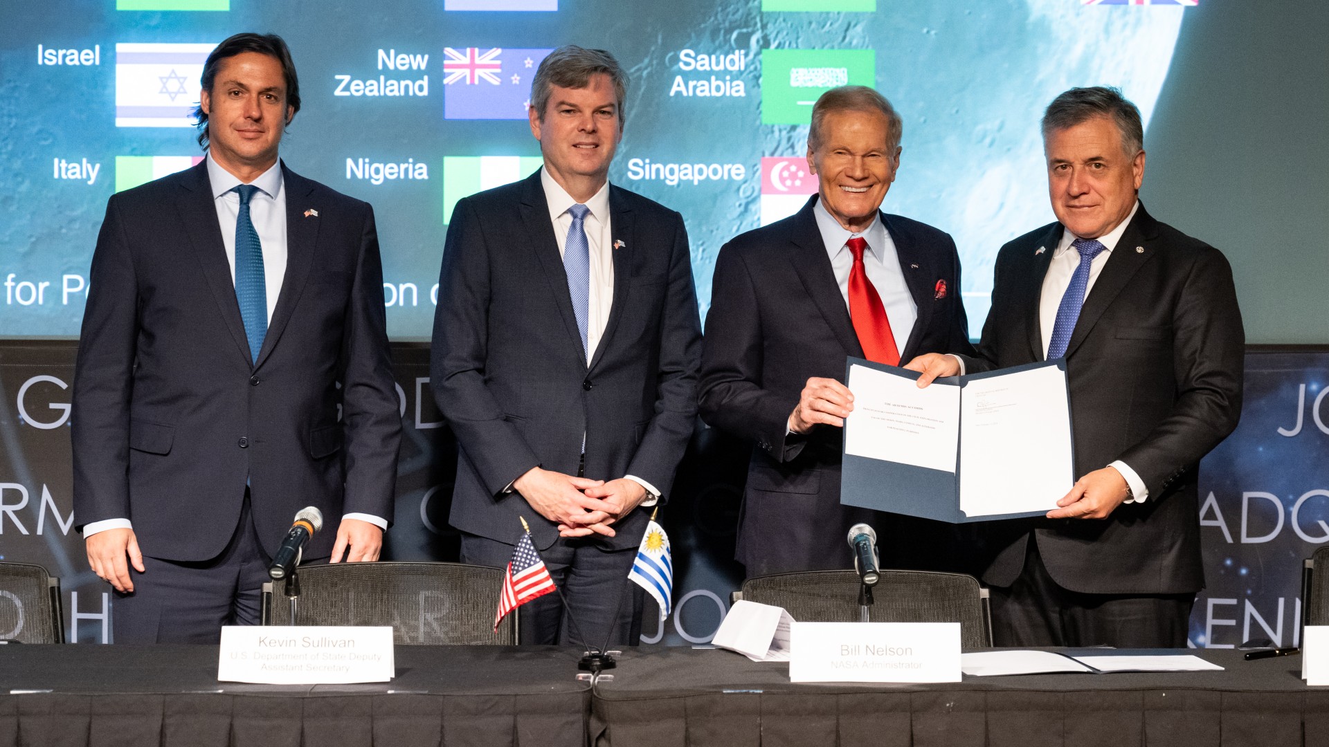 four men in suits smile and hold up a signed document for cameras to see