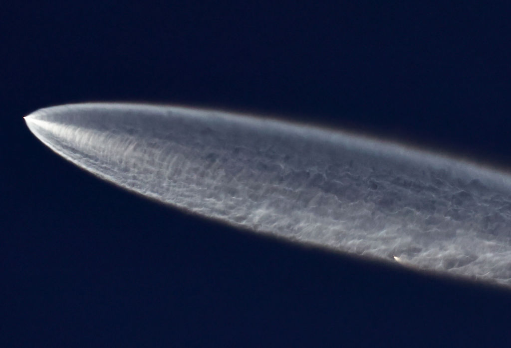 an expanding white plume extends from a small point of white light on the left, growing thicker in a stream across the darkening blue sky.