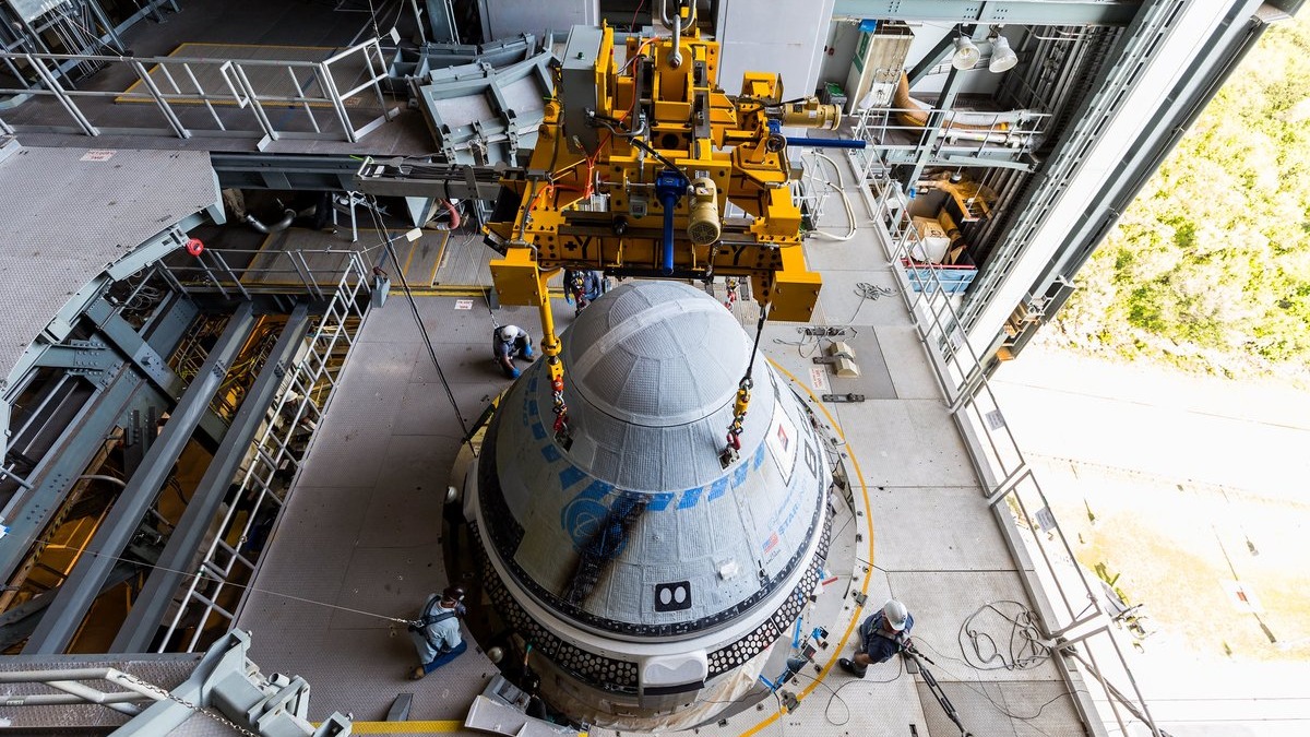 a cone-shaped spacecraft on a platform. an orange crane sits nearby. to the side is an open side of the building with grass and pavement visible